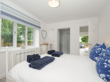 A bedroom with a white bed frame blue pillows and folded blue towels with a window and doors leading to a bathroom and another room at Otters Leap in Bideford
