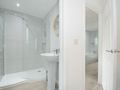 A bathroom with a shower, a pedestal sink, a round mirror, and a door leading to another bathroom with a bathtub at Otters Leap in Bideford