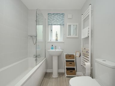 A bathroom with a bathtub shower combination a pedestal sink a toilet a window with a patterned blind and storage baskets at Otters Leap in Bideford