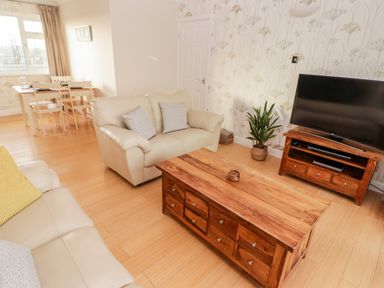 A living room with two beige sofas a wooden coffee table a wooden TV stand with a flat screen TV and a dining table with chairs by a window at 54 Croft Court in Tenby