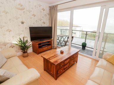 A living room with a wooden coffee table two beige sofas a television on a wooden stand a potted plant and a glass sliding door leading to a balcony at 54 Croft Court in Tenby