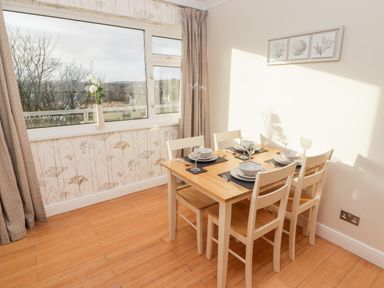 A dining area with a wooden table set for four near a window with floral wallpaper at 54 Croft Court in Tenby