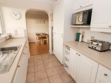 A kitchen with tiled floor white cupboards and countertops a microwave and toaster on the counter and a dining table set with plates in the background at 54 Croft Court in Tenby