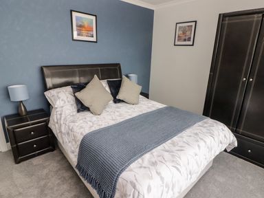 A bedroom with a bed with patterned bedding and cushions a bedside table with a lamp a wardrobe and framed pictures on the walls at 54 Croft Court in Tenby