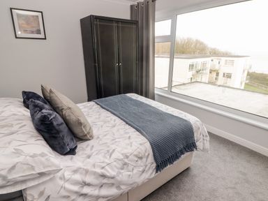 A bedroom with a bed, pillows, a blue throw, wardrobe, and a large window with a view of buildings and trees at 54 Croft Court in Tenby