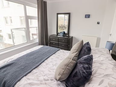 A bedroom with a bed with pillows and blanket beside a dresser with a mirror and a window with curtains at 54 Croft Court in Tenby