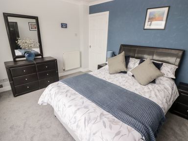 A bedroom with a bed dressed in patterned bedding and pillows a dresser with a vase and flowers and a mirror at 54 Croft Court in Tenby