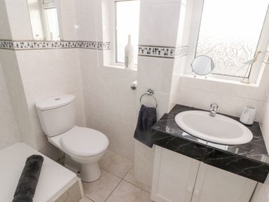 A bathroom with a toilet, sink with a black countertop, towel ring with a black towel, two windows with patterned glass, and a bathtub with a rolled black towel at 54 Croft Court in Tenby