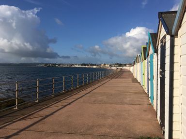 A seaside promenade with a row of colorful beach huts along the right side and a metal railing along the left side overlooking the sea at Little Rondeval in Totnes