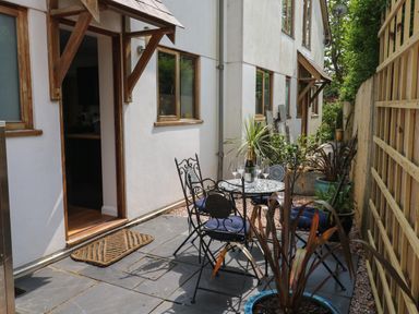 A small patio area with a round table and four chairs with blue cushions plants in pots along a fence at Little Rondeval in Totnes