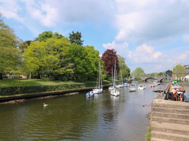 A river with boats moored along the bank trees on the opposite side and people sitting at tables on the riverside at Little Rondeval in Totnes