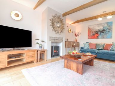 A living room with a blue sofa a wooden coffee table with fruit and chess set a wooden TV stand with a TV and a fireplace at Riviere Barton in Hayle