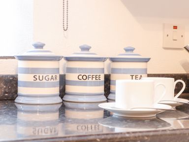 Three ceramic jars labeled sugar coffee and tea on a kitchen counter with two white cups and saucers in front at Riviere Barton in Hayle