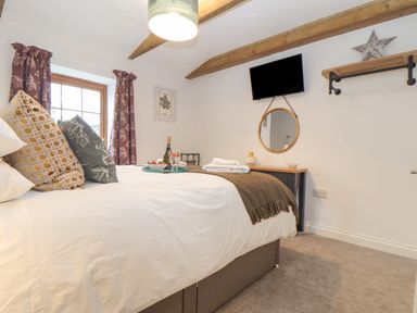 A bedroom with a bed pillows a blanket and a tray with a bottle and glasses a wall mounted tv a round mirror and wooden ceiling beams at Riviere Barton in Hayle