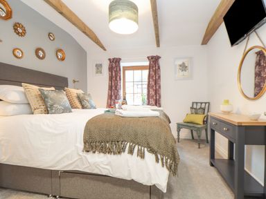A bedroom with a large bed cushions a gray headboard a window with curtains a chair a desk with decor and a wall-mounted television at Riviere Barton in Hayle