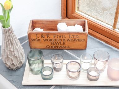 A collection of candles in glass holders on a wooden tray next to a wooden box with rolled towels and a vase with a yellow tulip on a stone windowsill at Riviere Barton in Hayle