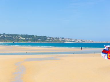 A sandy beach with people walking and kite surfers in the water near a coastal town at Riviere Barton in Hayle