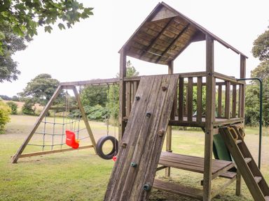 A wooden outdoor playset with a rope climbing wall swings slide and tire swing on grass at Riviere Barton in Hayle