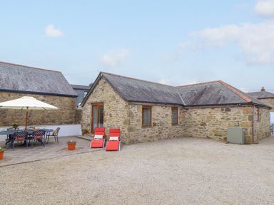 An outdoor area with stone buildings a patio table with chairs an umbrella and two red lounge chairs at Riviere Barton in Hayle