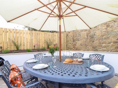 An outdoor patio with a round metal table set with plates glasses cutlery and a bread loaf under a large umbrella at Riviere Barton in Hayle