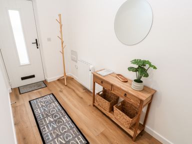 A hallway with a white door a wooden coat rack a wooden console table with baskets a plant and a circular mirror at Puffins Rest in Goldsithney