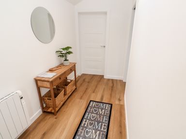 A hallway with wooden floor a wooden shelf with baskets a round mirror and a rug with home text at Puffins Rest in Goldsithney