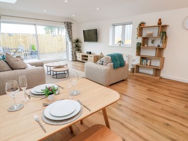 A dining table set with plates and glasses next to a living room with sofas a coffee table a TV and shelving at Puffins Rest in Goldsithney
