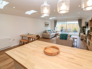 An open plan living room and dining area with a wooden table and chairs a beige sofa and armchair and patio doors leading outside at Puffins Rest in Goldsithney