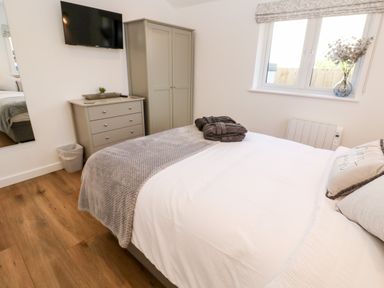 A bedroom with a bed covered in white and grey linens a chest of drawers a wardrobe a wall-mounted TV a window with a vase of flowers and wooden flooring at Puffins Rest in Goldsithney