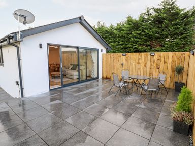 A patio with a glass table and four chairs next to a white building with sliding glass doors and a wooden fence at Puffins Rest in Goldsithney