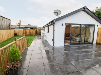 A patio with large tiles next to a white building with sliding glass doors and a fenced grassy area at Puffins Rest in Goldsithney