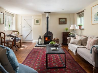 A living room with a wood stove a red patterned rug a beige sofa and a small dining table at Hawthorn Cottage at Collihole in Chagford