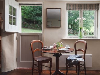 A small dining area with a round wooden table and two wooden chairs near a door and window with flowers on the table at Hawthorn Cottage at Collihole in Chagford