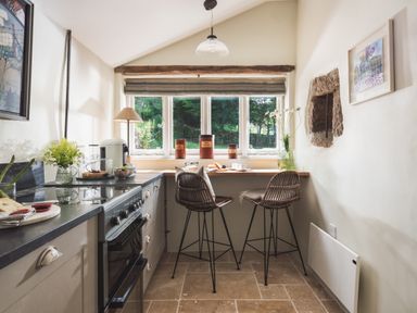 A kitchen with a stove countertop two high chairs and a window with jars and a lamp at Hawthorn Cottage at Collihole in Chagford
