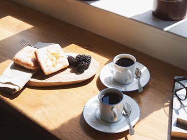 Two cups of coffee with spoons on saucers on a wooden table with pastries blackberries and a book with glasses at Hawthorn Cottage at Collihole in Chagford
