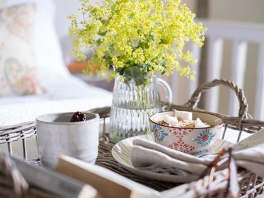 A wicker tray with a cup, bowl of cereal, cloth napkin, and a glass pitcher with yellow flowers at Hawthorn Cottage at Collihole in Chagford