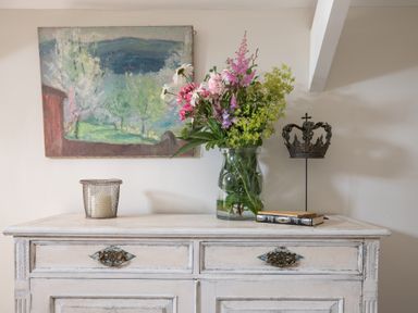 A white wooden dresser with a wire candle holder a vase of flowers two books and a metal crown sculpture with a landscape painting on the wall in the background at Hawthorn Cottage at Collihole in Chagford