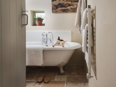 A bathroom with a clawfoot bathtub with a towel hanging over the side a wooden tray with bath items a small window with a potted plant and a towel rack with towels at Hawthorn Cottage at Collihole in Chagford