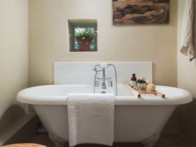 A bathroom with a white bathtub with a towel hanging over the edge and bath accessories on a wooden tray in the tub with a small window above the tub with a flower pot and a painting on the wall at Hawthorn Cottage at Collihole in Chagford