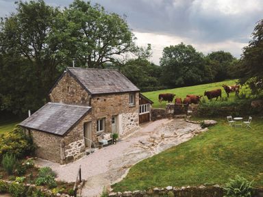 A stone cottage with a slate roof near a grassy field with cows and a small outdoor table and chairs set