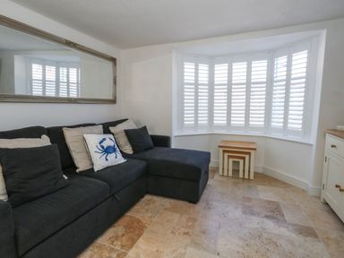 A living room with a dark sectional sofa and cushions near a window with white shutters and nested wooden tables at Belvedere Cottage in Weymouth