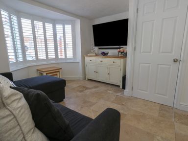 A living room with a sofa a bay window with white shutters a white cabinet with decorations and a wall mounted TV at Belvedere Cottage in Weymouth