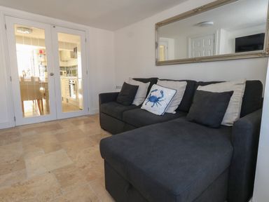 A living room with a dark sofa with cushions and glass doors leading to a kitchen at Belvedere Cottage in Weymouth