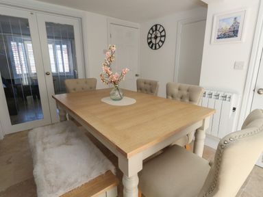 A dining room with a wooden table surrounded by beige upholstered chairs and a bench with a white fur cover at Belvedere Cottage in Weymouth