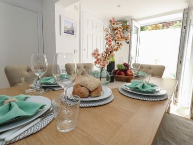 A dining table set with plates napkins glasses bread and fruit bowl in a room with beige chairs at Belvedere Cottage in Weymouth