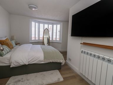 A bedroom with a bed pillows and blanket a standing mirror window shutters a wall-mounted television radiator and a small bedside table at Belvedere Cottage in Weymouth