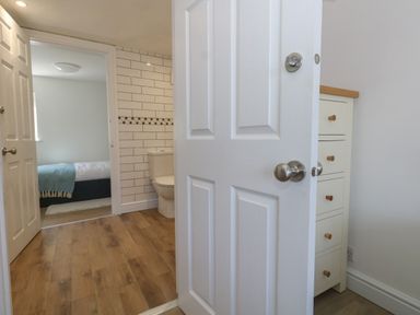 A hallway with wooden floor leading to a bathroom with a toilet and a bedroom with a bed covered by a blue blanket at Belvedere Cottage in Weymouth