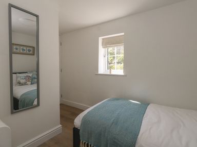 A small bedroom with a single bed covered with a white duvet and blue throw next to a window and a wall mirror at Belvedere Cottage in Weymouth