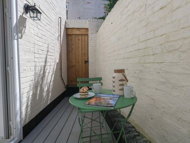 A small outdoor patio with a green table and chair set two mugs a muffin a lantern and a magazine at Belvedere Cottage in Weymouth