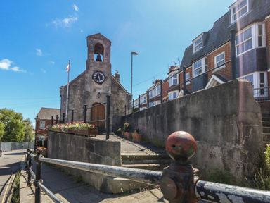 A street view showing a stone clock tower with a bell and nearby red brick houses at Belvedere Cottage in Weymouth